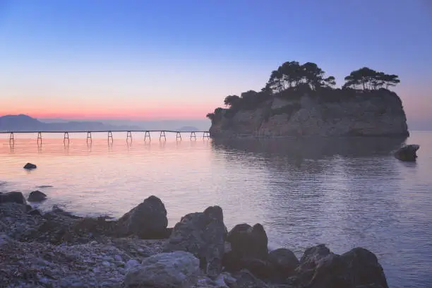 Photo of Small tropical island with wooden hanging bridge from Zante Greek island to it. Panoramic morning dawn scenery. Aegean sea. Zante Zakinthos is popular Greek resort. Opaque artistic processing.