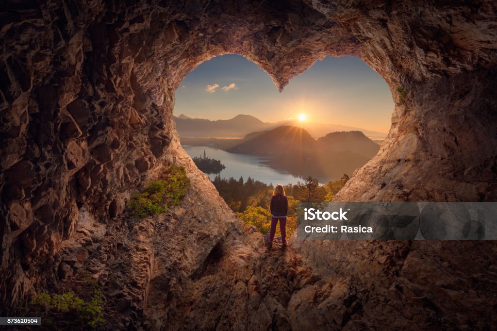 Young woman in heart shape cave towards the idyllic sunrise Lone woman enjoying in beautiful mountain nature, celebrating freedom and standing on edge of the cliff against the rising sun. Valentines day concept. Heart Shape Stock Photo