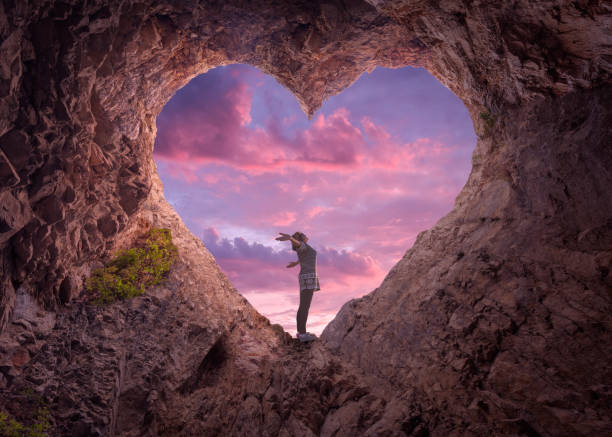 Young woman in heart shape cave towards the beautiful sky Happy woman enjoying in idyllic mountain nature, celebrating freedom and rising her arms while standing toward the setting sun. Valentines day concept. attached stock pictures, royalty-free photos & images