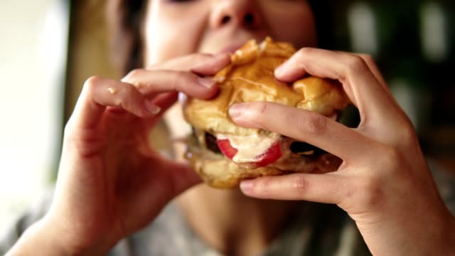 Closeup view of young woman biting big tasty juicy burger in cafe. Slowmotion shot