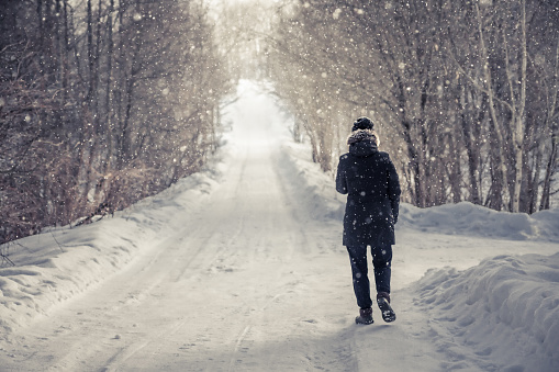 Single woman walking on snowy winter road among trees  alley with light at the end of the way in cold winter  day during snowfall  with copy space