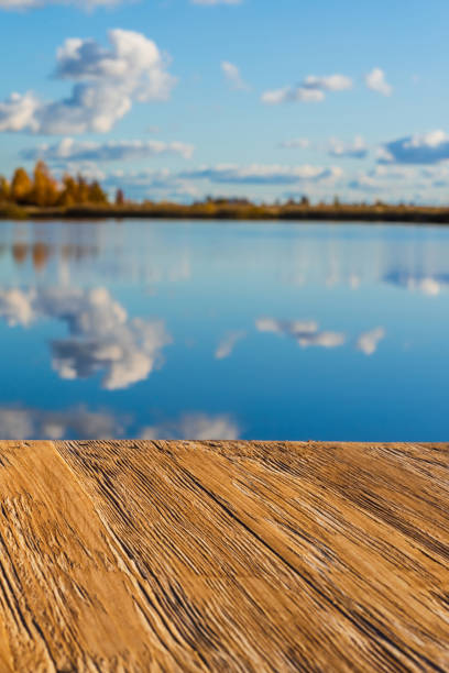 istock Empty rustic wood table top on  blurred colorful autumn landscape background with fall forest and blue sky with cloudscape reflection on the lake. Can montage or display your products 873701596