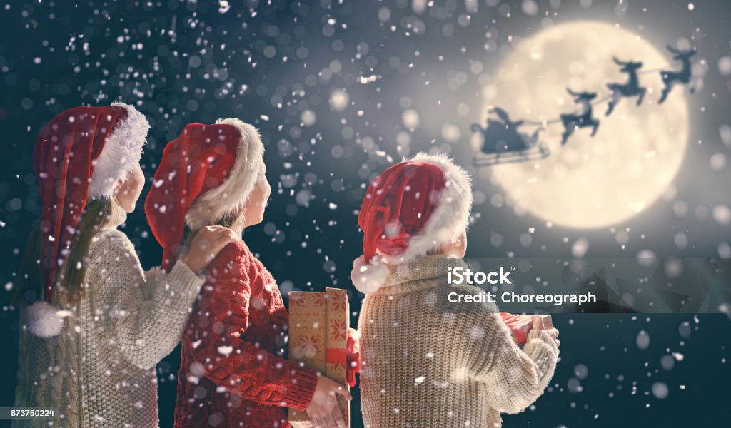 children with xmas presents Merry Christmas and happy holidays! Cute little children with xmas presents. Santa Claus flying in his sleigh against moon sky. Kids enjoying the holiday with gifts on dark background. Christmas Stock Photo