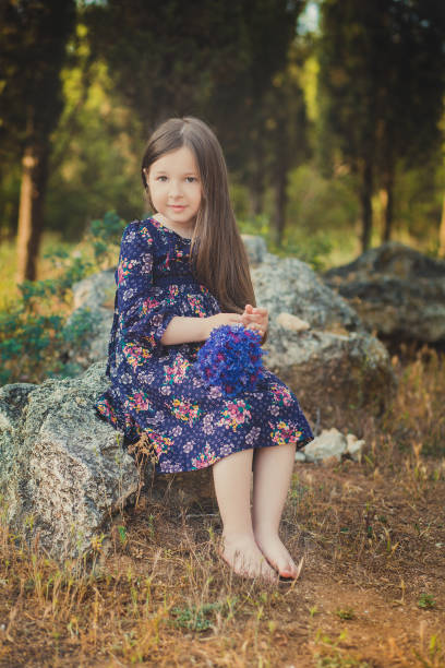 mignon bébé fille avec des cheveux brunette et portrait de brun yeux bleu profond violet fleurs sauvages aux pieds nus, robe élégante fleur colorée et en regardant vers la caméra avec brillant visage - child playing dressing up imagination photos et images de collection