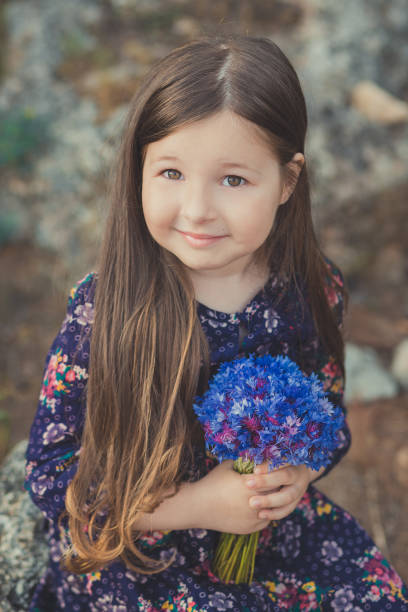 mignon bébé fille avec des cheveux brunette et portrait de brun yeux bleu profond violet fleurs sauvages aux pieds nus, robe élégante fleur colorée et en regardant vers la caméra avec brillant visage - child playing dressing up imagination photos et images de collection