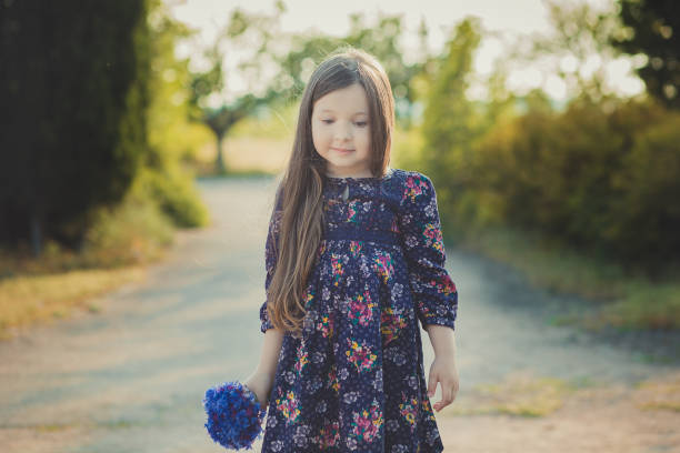 mignon bébé fille avec des cheveux brunette et portrait de brun yeux bleu profond violet fleurs sauvages aux pieds nus, robe élégante fleur colorée et en regardant vers la caméra avec brillant visage - child playing dressing up imagination photos et images de collection