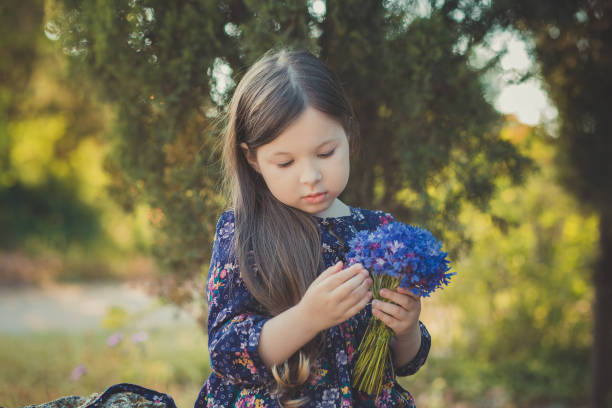 mignon bébé fille avec des cheveux brunette et portrait de brun yeux bleu profond violet fleurs sauvages aux pieds nus, robe élégante fleur colorée et en regardant vers la caméra avec brillant visage - child playing dressing up imagination photos et images de collection