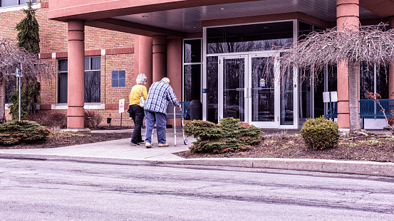 An elderly senior adult man using an orthopedic medical equipment walker to support himself as he walks is entering a medical office building near Rochester, New York State, USA. His senior adult woman home caregiver daughter is accompanying him to help with his doctor's appointment.