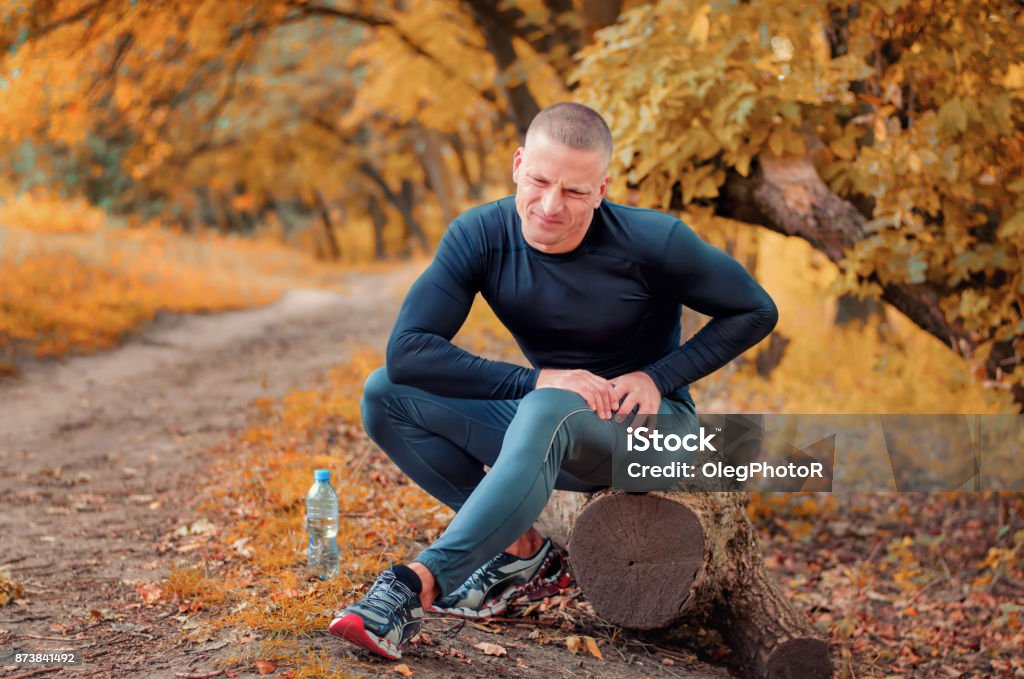 A young athletic  jogger feels a strong pain in the muscle after cramping. A young athletic  jogger in black tight fitting sportswears and  sneakers sits on a log, feels a strong pain in the muscle after cramping on a autumnall forest background. Pain Stock Photo
