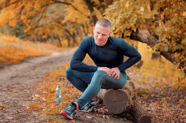 A young athletic  jogger feels a strong pain in the muscle after cramping. A young athletic  jogger in black tight fitting sportswears and  sneakers sits on a log, feels a strong pain in the muscle after cramping on a autumnall forest background. carpet runner stock pictures, royalty-free photos & images