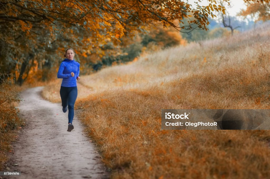 young slim athleic jogger woman runs along the path  on the  beautiful  autumnal forest. young slim athleic jogger in a black sports leggins and and blue jacket  runs along the path  on the  beautiful  autumnal forest. Photo show  active healthy lifestyle. Autumn Stock Photo