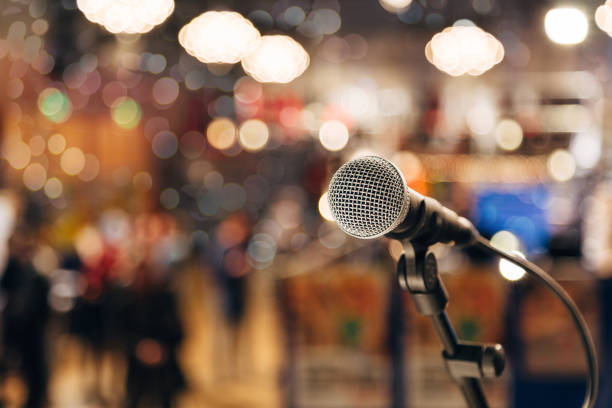 istock Microphone on a stand ready for live music performance or karaoke night with soft bokeh lights and people silhouettes in the background. Concept for musical singing event, having a good time. 873861320