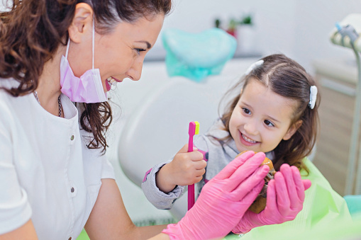 Smiling girl sitting in dentist chair with her pediatric dentist showing her teeth model and teaching her how to brush teeth. Dentistry, oral hygiene concept