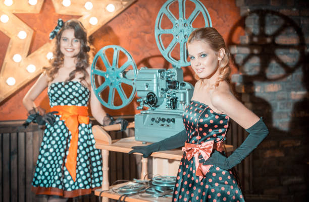 The two beautiful girls (with blonde hair and with brown hair) dressed in a vintage polka dot dresses are standing near the old film projector. The girls are smiling looking at the camera. Studio shooting
