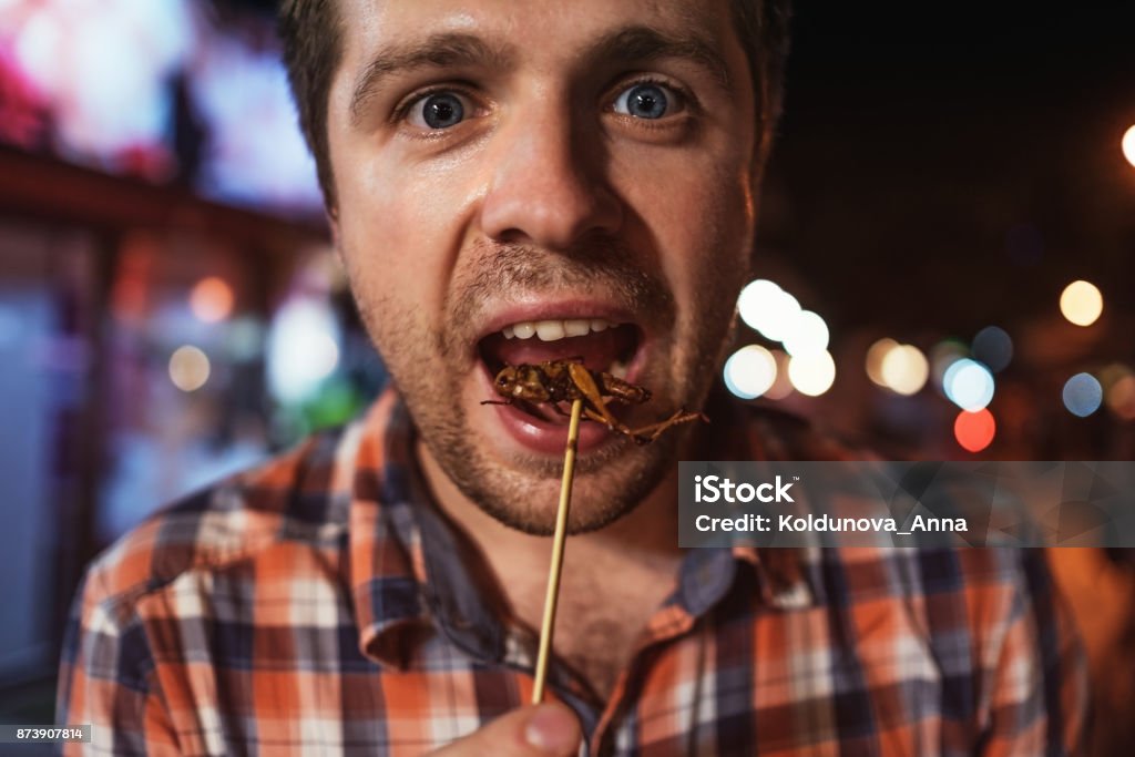 Caucasian young male eating cricket at night market in Thailand. Caucasian young male eating cricket at night market in Thailand. Eating insect concept Eating Stock Photo