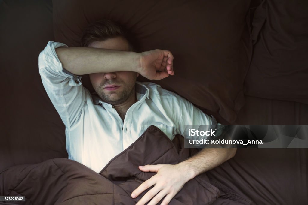 Portrait of a young caucasian man from above sleeping in a dark bed. Portrait of a young caucasian man from above sleeping in a dark bed. He is tired after work and sleep in white shirt Above Stock Photo