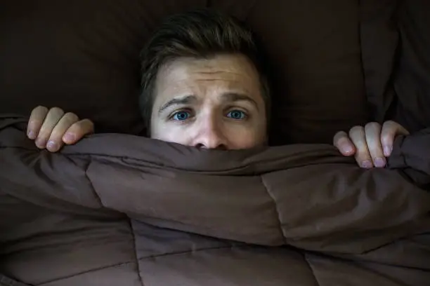 Photo of Caucasian young man hiding in bed under the blanket at home.