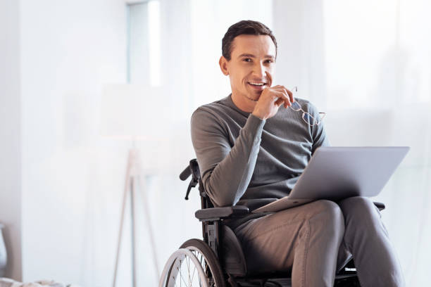 Positive delighted disabled man looking forward Having secret. Enigmatical male keeping smile on his face and holding glasses in right hand while sitting on the wheelchair wheelchair photos stock pictures, royalty-free photos & images