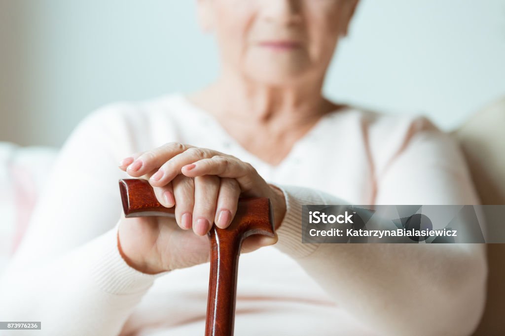 Elder holds hands on walking stick Elder holds hands on walking stick while sitting on white sofa in nursing home Parkinson's Disease Stock Photo