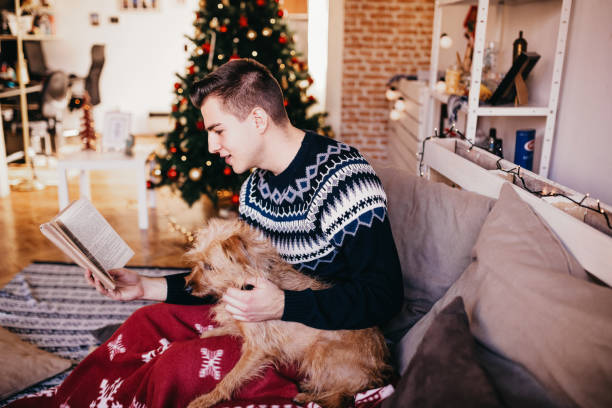 istock Young man is sitting on a sofa and reading a book while cuddling with his dog, Christmas decoration 874016898