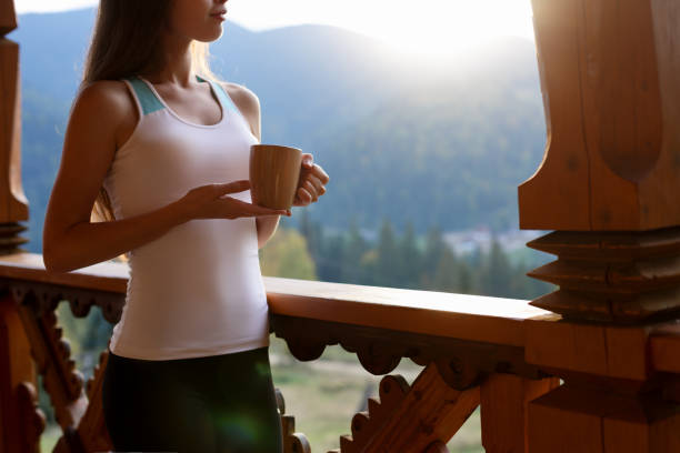 Slim caucasian woman holds cup of tea in her hands at mountain resort. Sports girl with hot coffee mug at wooden balcony of country house. Forest and mountains on background. Diet concept. stock photo