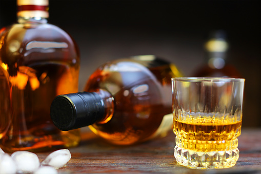 Close-up of a glass of whiskey standing on a rustic wooden table with ice cubes laying next to it. Several generic looking whiskey bottles can be seen in the background.