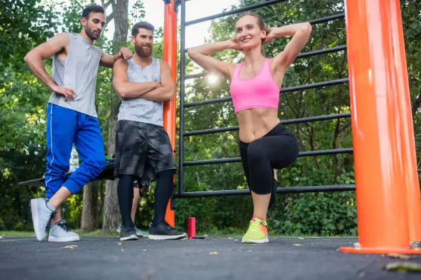Full length of a ravishing young woman smiling while doing Bulgarian squats during lower-body workout routine in the admiration of her male friends outdoors