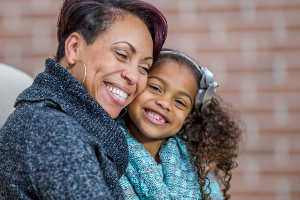 A mother and daughter of African descent are indoors in their living room. They are wearing warm sweaters. They are both smiling while sitting on a sofa together.