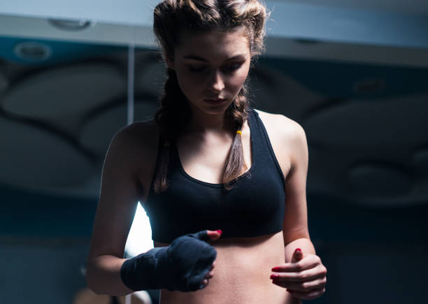 Young fighter boxer girl putting on hand bandage Young beautiful fighter boxer girl putting on hand bandage before training win dark gym. Low key image. boxercise stock pictures, royalty-free photos & images