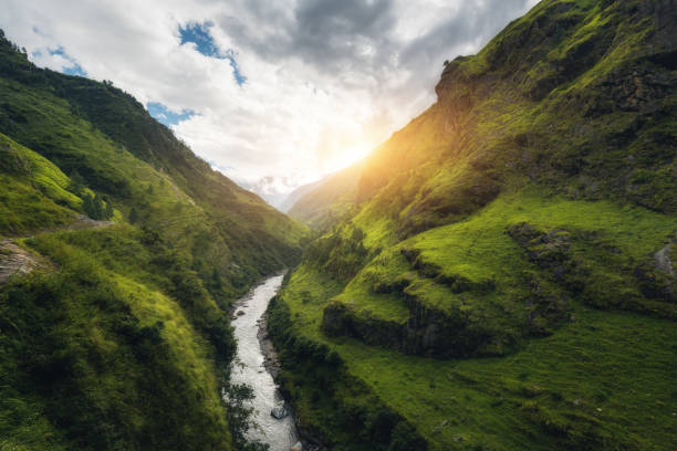istock View with amazing Himalayan mountains covered green grass, river, meadows and forest, blue sky with clouds, sun and stones in water in autumn in Nepal at sunrise. Mountain valley. Travel in Himalayas 874176278