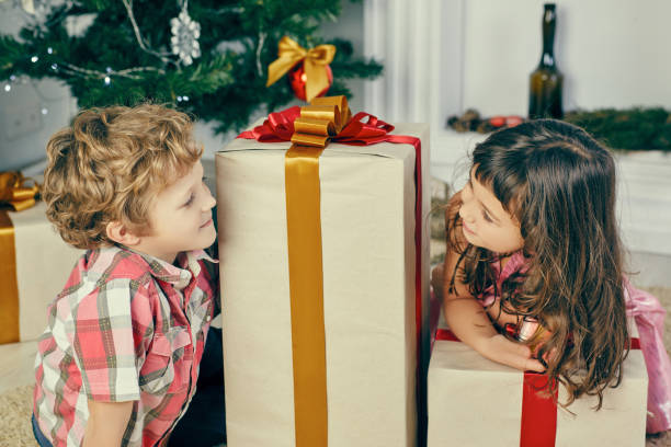 little girl peeks out from behind big christmas gift box and looks at boy. - christmas child little boys peeking imagens e fotografias de stock