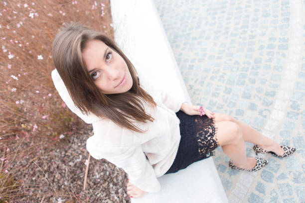 Young latin woman crouching in a park beside a tree, wearing a stylish white shirt, black vest, plaid skirt, and black boots while confidently looking at the camera