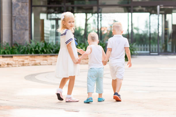Little girl and two boys holding hands and walking outdoors with building in background. Girl is turning round and looking at camera. Back view.