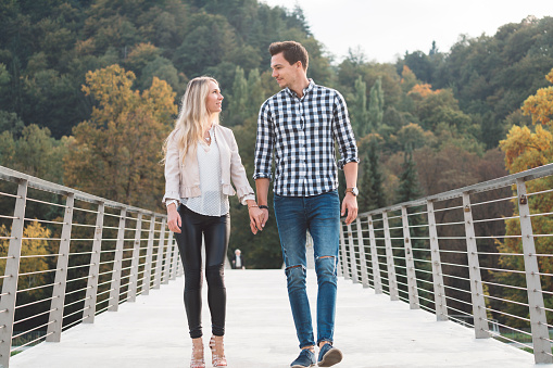 Young couple walking across the bridge, holding hands, having fun, wearing modern clothes, looking at each other, being in love.