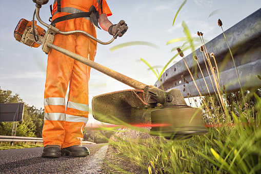 Professional road worker with bright clothing trimming grass underneath a guard railing.
