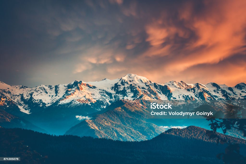 Sunset evening view over the snowy mountain range Beautiful colorful sunset over the snowy mountain range and pine tree forest. Nature landscape. Dramatic overcast sky with orange clouds. Main Caucasian ridge, Svaneti, Georgia. Retro toning filter Autumn Stock Photo