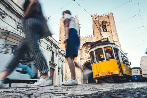 Traditional yellow tram driving on R. Augusto Rosa street towards Lisbon Cathedral. Tourists are crossing the street.