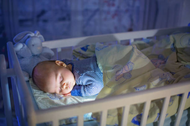 Adorable newborn baby boy, sleeping in crib at night. Little boy in blue striped pajamas taking a nap in dark room, christmas decoration in the room, winter time, snowing outdoors