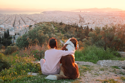Young woman hugs her dog as they sit in a field.Athens,Greece.Real warm light from sunset.