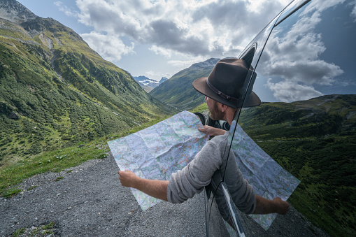 Young man in car on mountain road looks at map for directions. Mountain landscape in Summer, shot in Graubunden Canton, Switzerland.