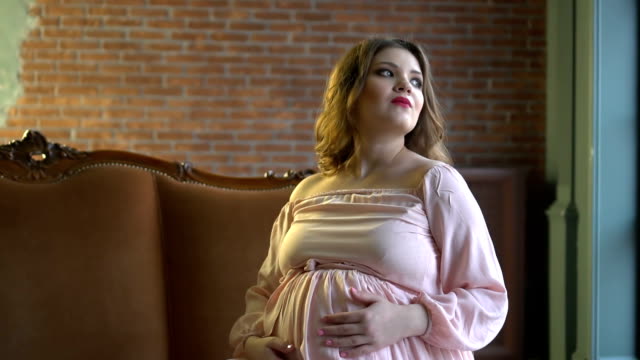 Very beautiful young pregnant woman sitting on a couch and holds her hands on her stomach.