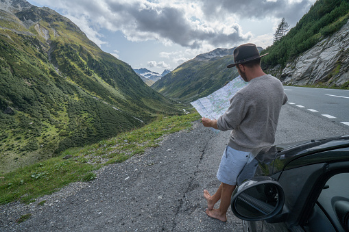 Young man in car on mountain road looks at map for directions. Mountain landscape in Summer, shot in Graubunden Canton, Switzerland.