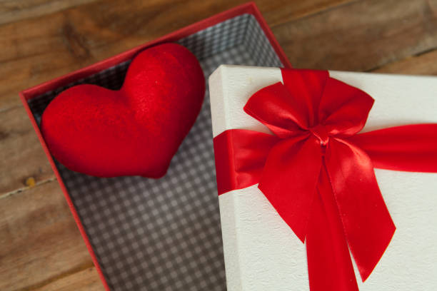 White gift box and red ribbon and mini heart inside on wood background with space.