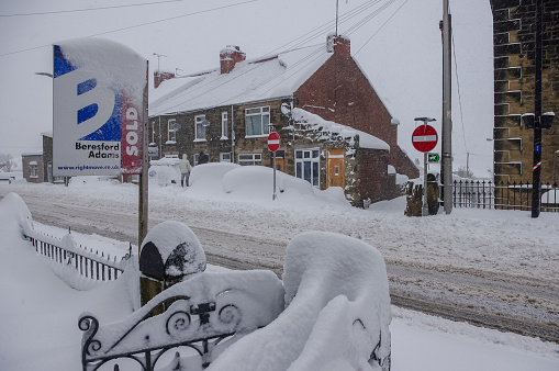Wrexham, UK - March 22, 2013: Street scene in Wales during heavy snow and blizzard conditions. Deep snow covering everything including cars, pathways, road, and for sale sign. People viewing damage.