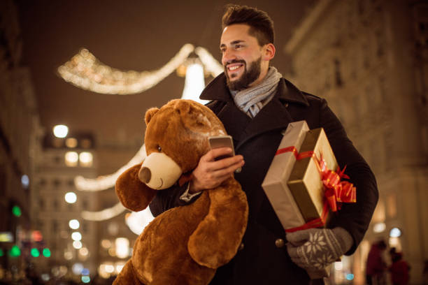 Can't wait to see her face Young man holding Christmas gifts and big Teddy bear. Checking his smart phone. Wearing warm clothing. Vienna, Austria. man with teddy bear stock pictures, royalty-free photos & images