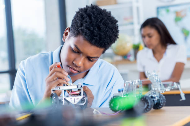 Focused teenage boy builds robot at school Teenage boy concentrates as he builds a robotic vehicle during engineering class. He is using a tool on the robot. A female student is working in the background. independent school education stock pictures, royalty-free photos & images