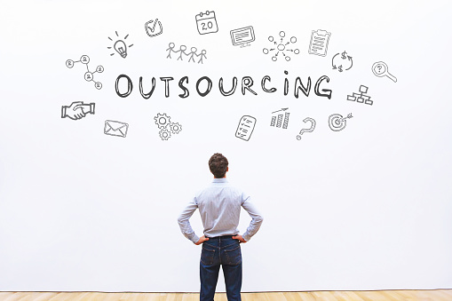 A man stands with his back to the viewer, facing a white wall decorated with various doodles and icons related to "outsourcing," including a handshake, charts, and light bulbs, creating a thematic visual representation of the concept.