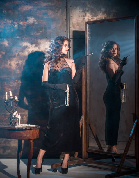 Attractive Woman before Night Party Beautiful woman with long brown hair is dressed in black evening dress. She is standing near the full-length mirror and holding the cigarette mouthpiece in hand. The young woman is looking on her reflection in the mirror over her shoulder. Studio shooting at dark woman full length mirror stock pictures, royalty-free photos & images