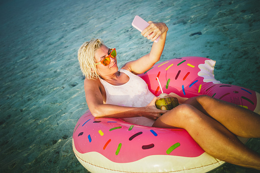One young and attractive woman is floating in inflatable rubber. Rubber have shape of doughnut which is missing one part, something like one bite and it's colored in pink and yellow. This sexy woman is taking selfie while enjoying on this heavenly island. She is holding coconut fruit with coconut water in it and straw. Short blonde hair is wet and her white swimsuit so she was swimming a little before. Water is crystal clear and turquoise colored. It's sunny summer day, perfect for enjoyment and relaxing in the ocean.