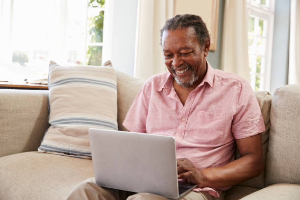 último homem sentado no sofá usando o laptop em casa juntos - idoso na internet - fotografias e filmes do acervo